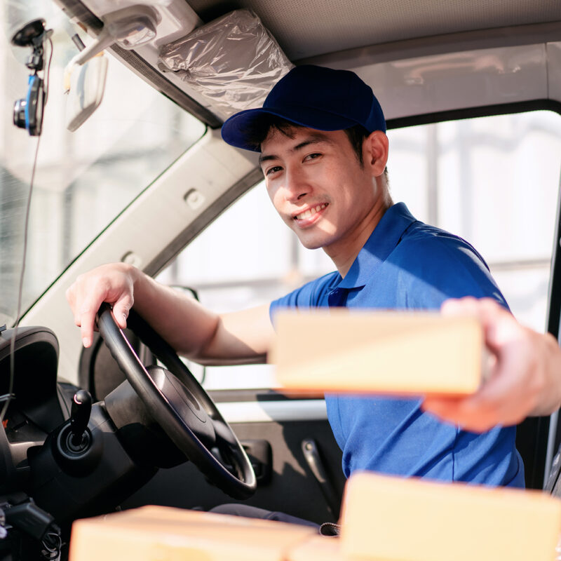 A man in a blue shirt is driving a truck and smiling. He is holding a box in his hand